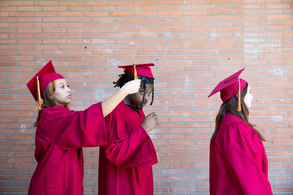 Two Hispanic female students in graduation cap and gown adjusting the mortarboard of a Black male student in preparation for a graduation ceremony on a college campus.
