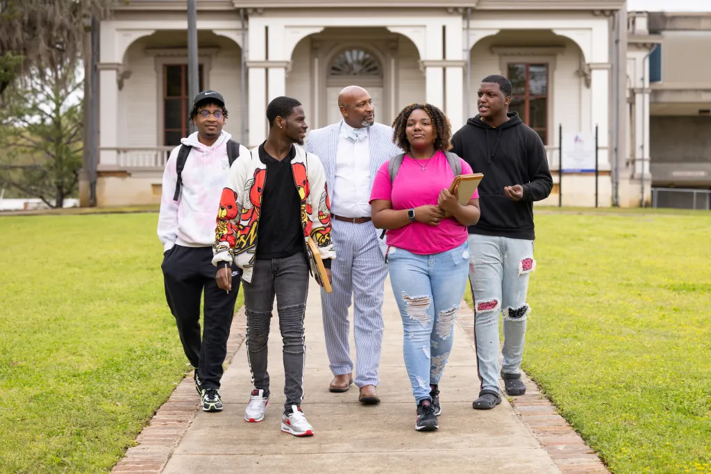 A group of four students and a professor walk together on a campus pathway, talking and smiling in front of a historic building.