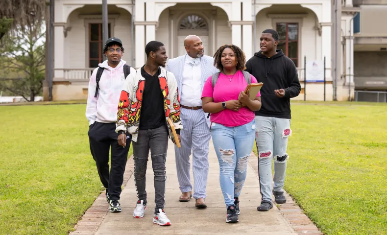 A group of four students and a professor walk together on a campus pathway, talking and smiling in front of a historic building.