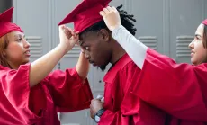 Two graduates in red gowns help adjust the cap of another graduate before a ceremony.
