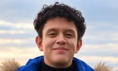 Adam Acosta smiling gently at the camera, wearing a bright blue winter jacket with a fur-trimmed hood against a softly blurred cloudy sky.