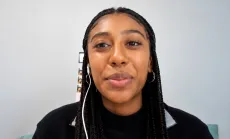 Mya Vickers looking at the camera with a subtle smile, wearing a black top and hoop earrings, with long braids and white earbuds.