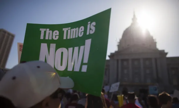 Green sign with white letters stating "The Time Is Now!" in front of city hall