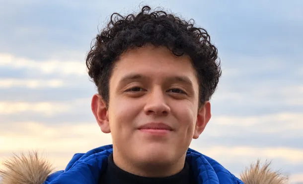 Adam Acosta smiling gently at the camera, wearing a bright blue winter jacket with a fur-trimmed hood against a softly blurred cloudy sky.