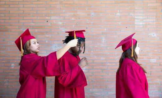 Two Hispanic female students in graduation cap and gown adjusting the mortarboard of a Black male student in preparation for a graduation ceremony on a college campus.