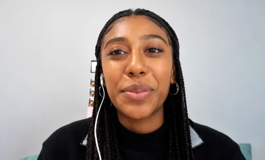 Mya Vickers looking at the camera with a subtle smile, wearing a black top and hoop earrings, with long braids and white earbuds.